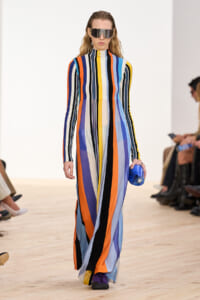 Model walking a runway in a floor-length, multicolored striped dress with a high collar and oversized sunglasses, holding a blue clutch.