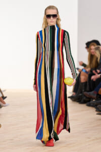 Model walking a runway in a floor-length, multicolored striped dress with long sleeves and a high neck; she wears oversized sunglasses and coral shoes, and holds a yellow fruit accessory.
