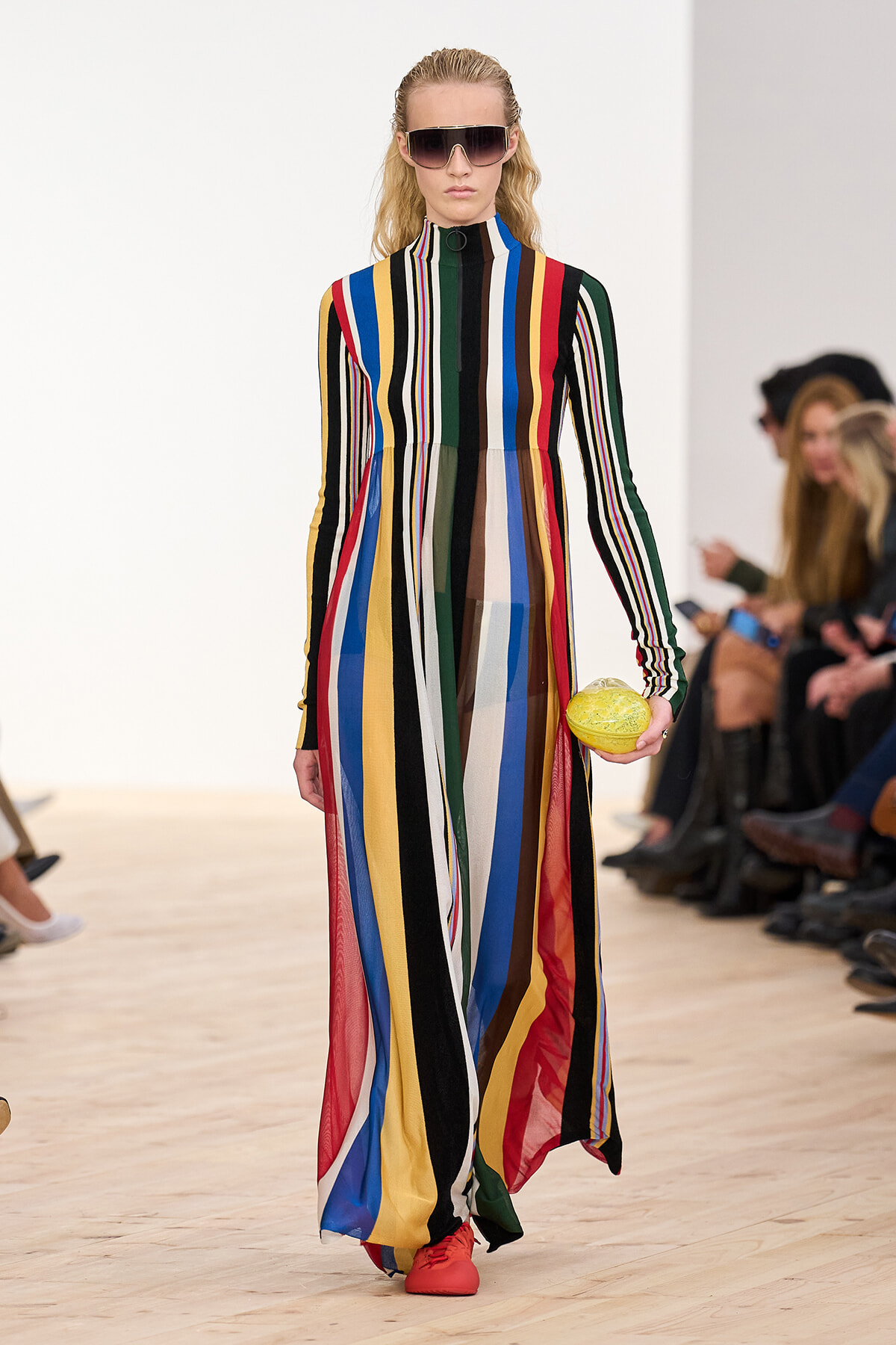 Model walking a runway in a floor-length, multicolored striped dress with long sleeves and a high neck; she wears oversized sunglasses and coral shoes, and holds a yellow fruit accessory.