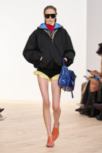 Female model on a runway wearing an oversized black windbreaker, yellow shorts, orange shoes, and a bright blue handbag, with large sunglasses.