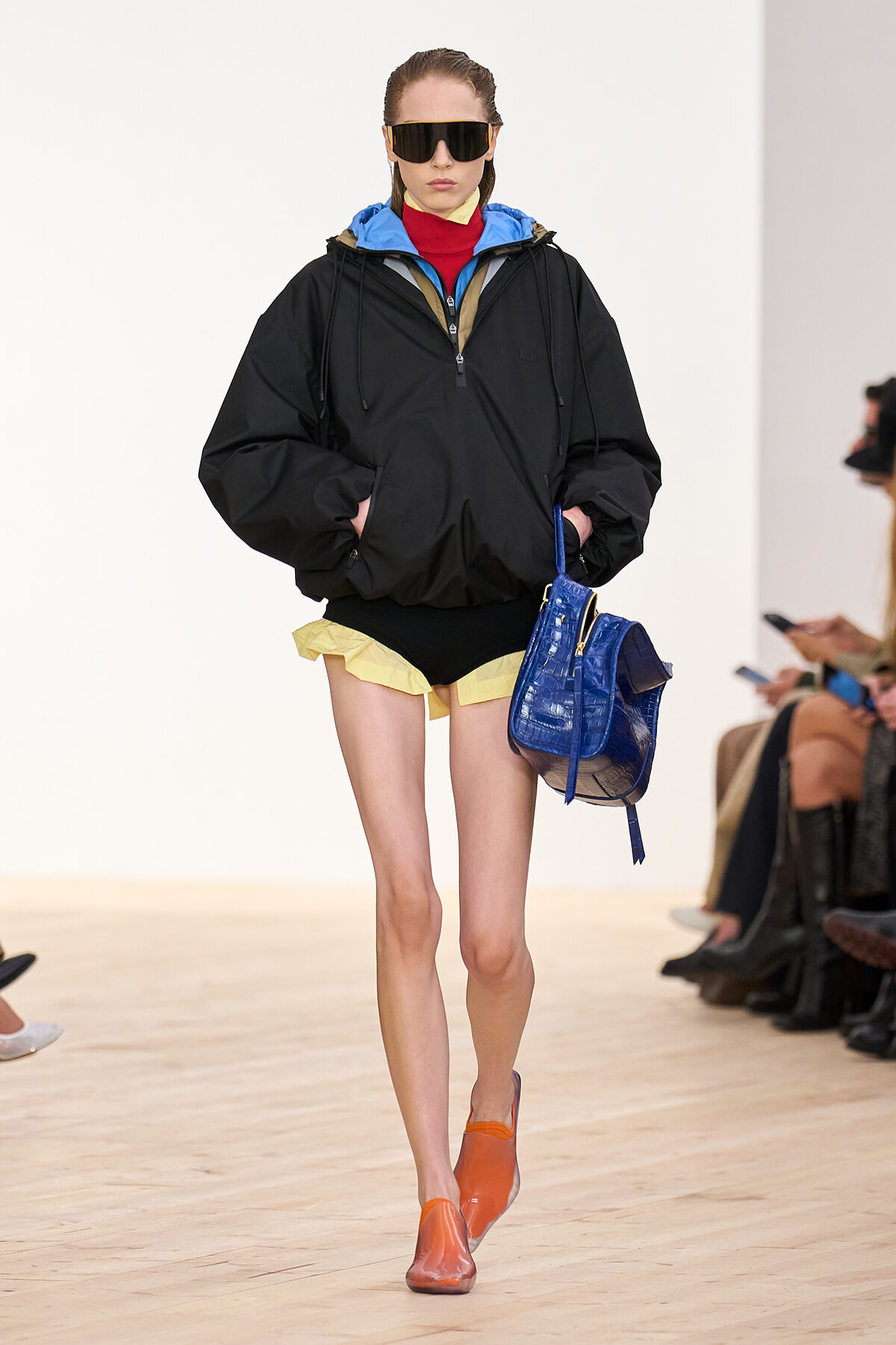 Female model on a runway wearing an oversized black windbreaker, yellow shorts, orange shoes, and a bright blue handbag, with large sunglasses.