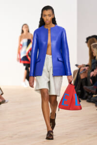Model walks the runway in a royal blue blazer over cream textured shorts, carrying a red bag with blue letters.