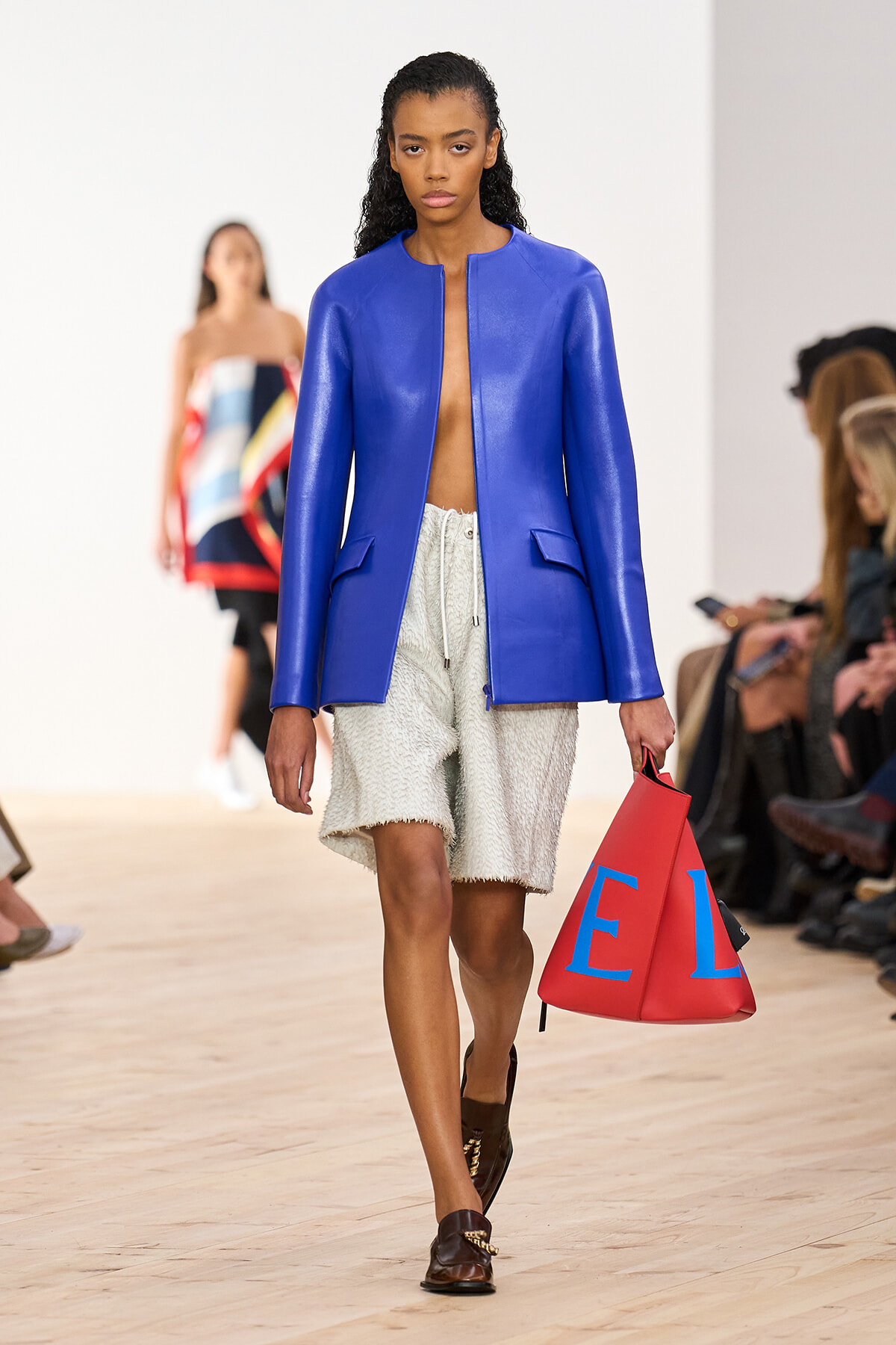Model walks the runway in a royal blue blazer over cream textured shorts, carrying a red bag with blue letters.