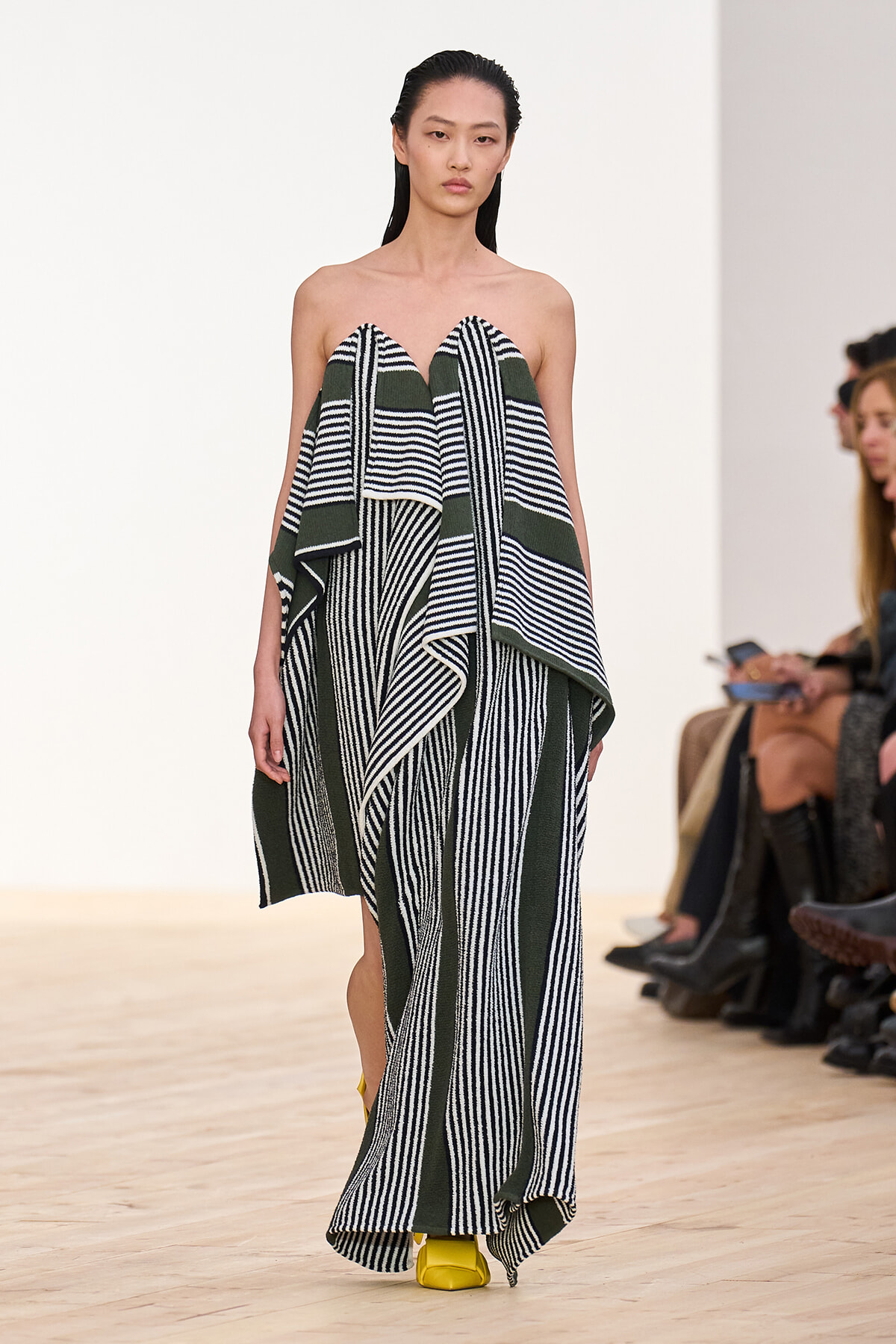 Model on runway wearing a strapless green and black striped draped gown with long, flowing panels.