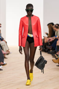 Model on the runway wearing a coral red leather blazer over black high-cut shorts, bright yellow loafers, oversized sunglasses, and carrying a black handbag.