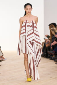 Runway model wearing a white and burgundy striped strapless draped dress with yellow flats.