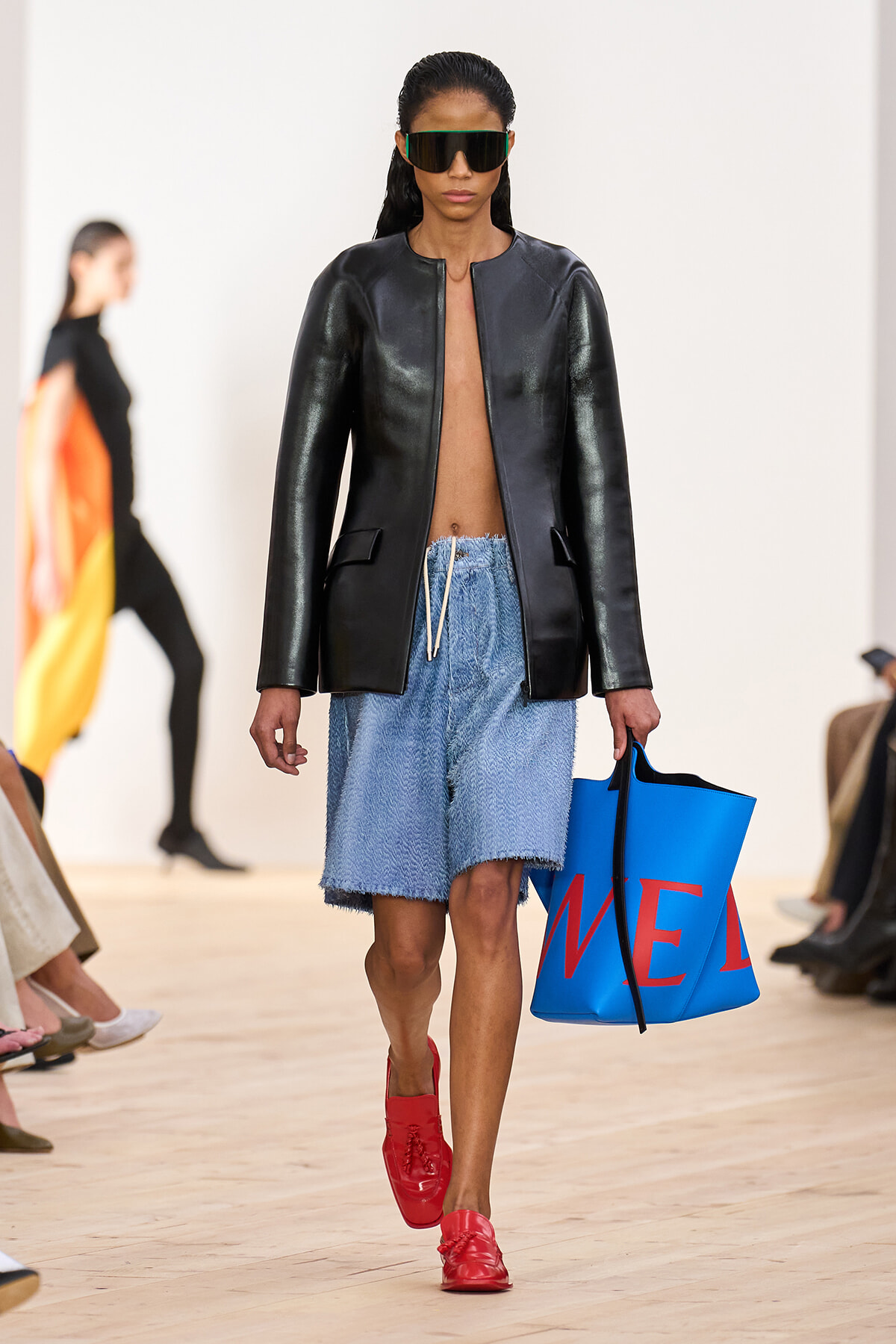Model walking in a black leather jacket, light-blue denim shorts, red loafers, oversized sunglasses, carrying a bright blue tote with red letters on a runway.