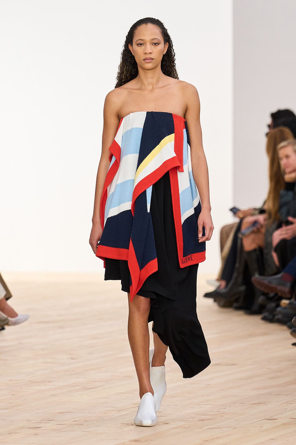 Model on a runway wearing a strapless, color-block striped wrap with red borders over a black dress; white ankle boots visible in the foreground.