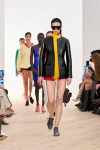 Model walks a fashion runway wearing a black leather jacket over a bright yellow outfit with red shorts; large sunglasses and slicked-back hair.