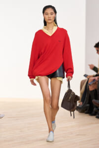 Model walking a runway in an oversized red sweater layered over a beige collar, paired with black shorts and a brown handbag.