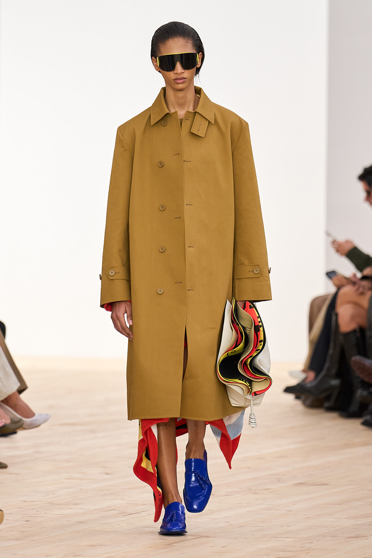 Female model on a runway wearing a camel oversized coat, large black sunglasses, electric blue loafers, and holding a multicolored sculptural bag.