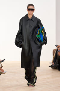 Model on a runway wearing an oversized black leather coat and large sunglasses, carrying a stack of bright blue, green, and yellow fabrics.