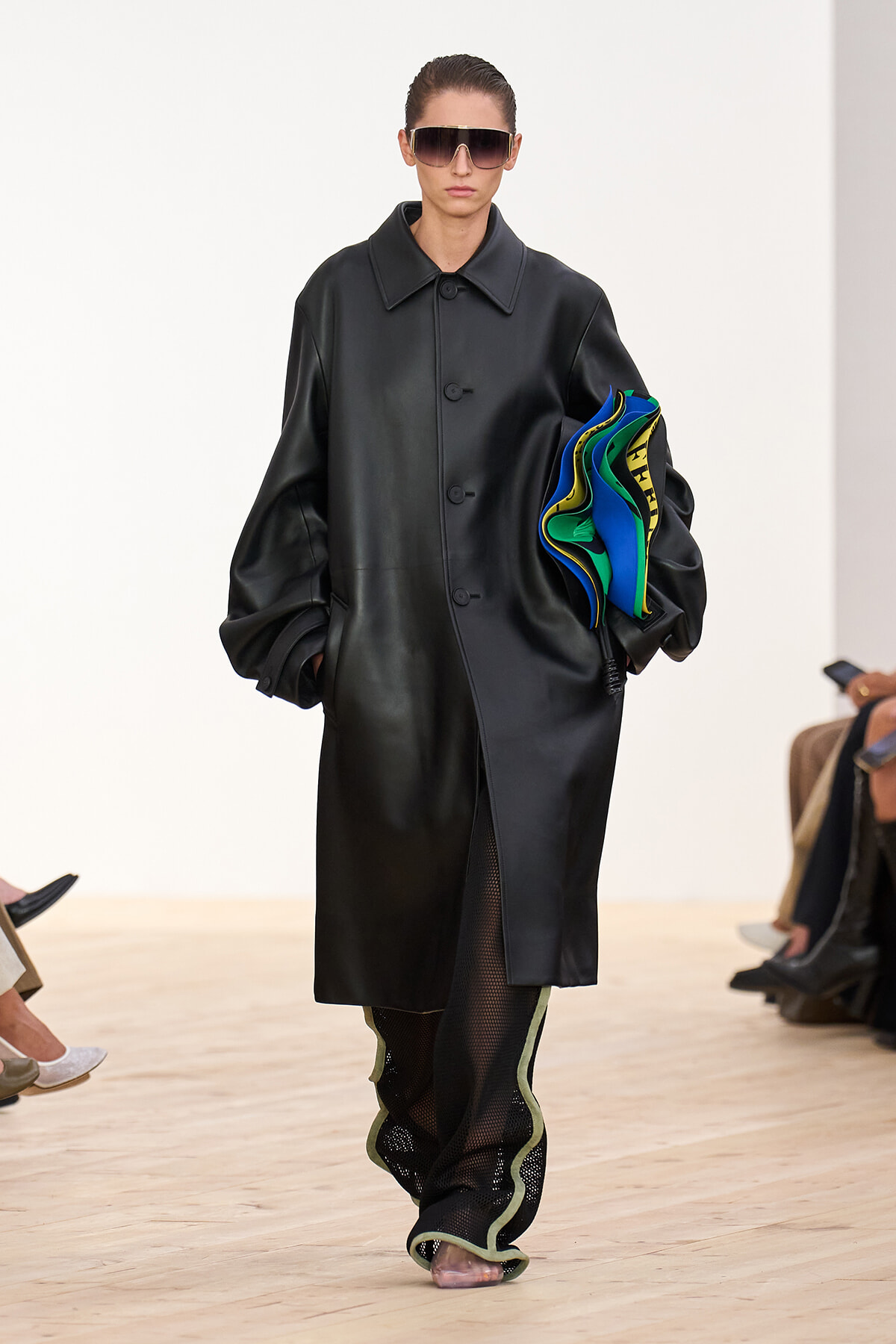 Model on a runway wearing an oversized black leather coat and large sunglasses, carrying a stack of bright blue, green, and yellow fabrics.