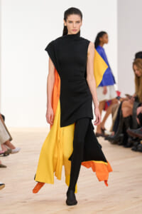 Female runway model in a black asymmetrical sleeveless dress with a yellow and orange cape-like overlay flowing behind her on a wooden catwalk.