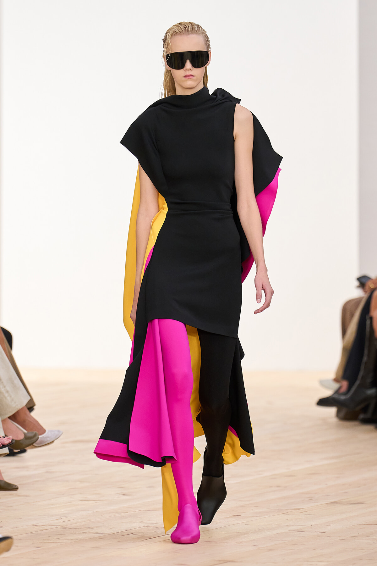 Runway model in a black dress with magenta and yellow cape panels and oversized sunglasses.