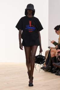 Black model on a runway wearing an oversized black Loewe T‑shirt with a red/blue logo, a wide-brim hat, and a black sculptural handbag空