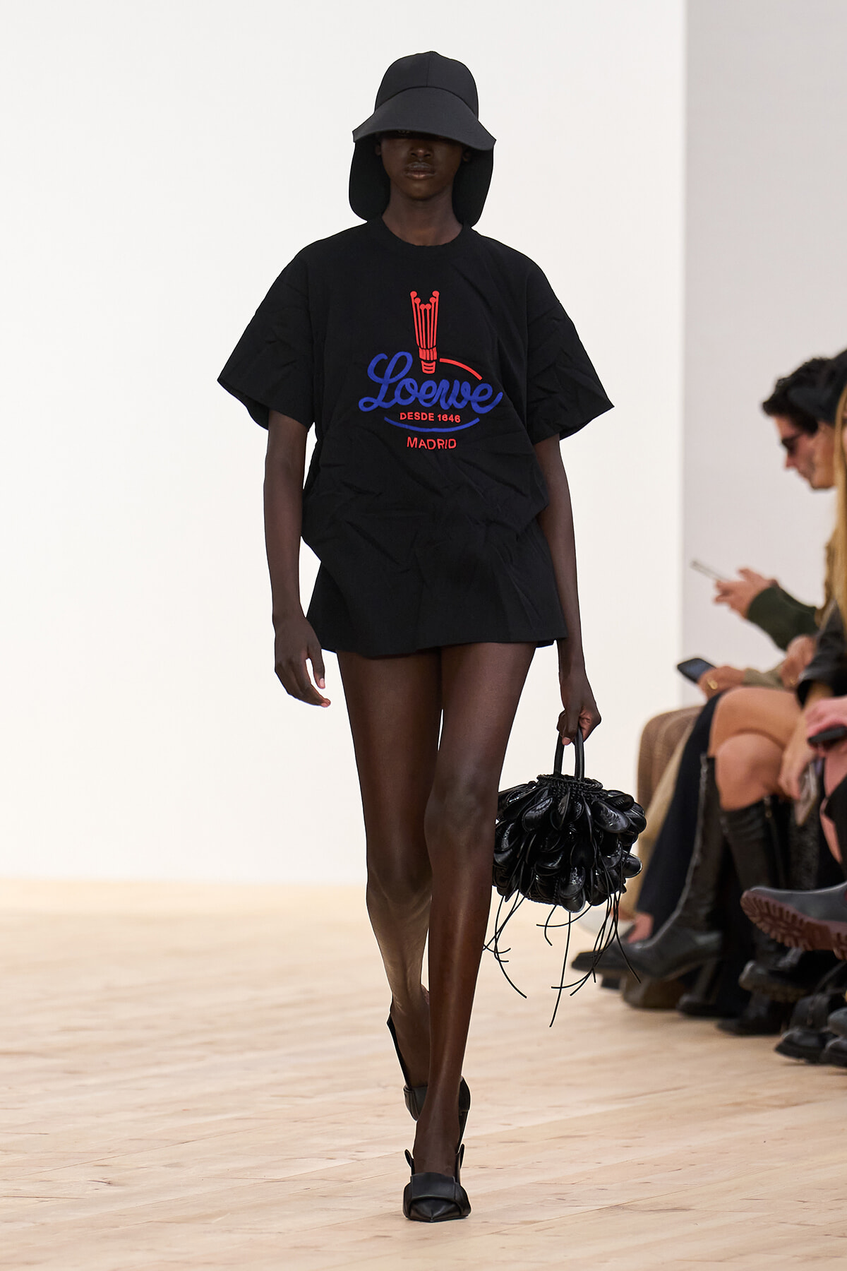 Black model on a runway wearing an oversized black Loewe T‑shirt with a red/blue logo, a wide-brim hat, and a black sculptural handbag空