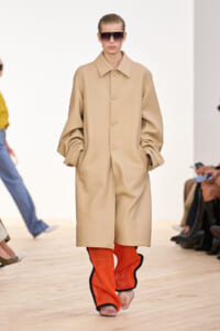 Model walking a runway in an oversized beige coat paired with bright orange mesh pants and large sunglasses.