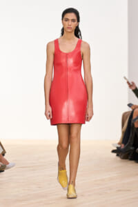 Model walking a runway in a sleeveless red leather mini dress with a centered vertical seam, paired with yellow slip-on shoes.