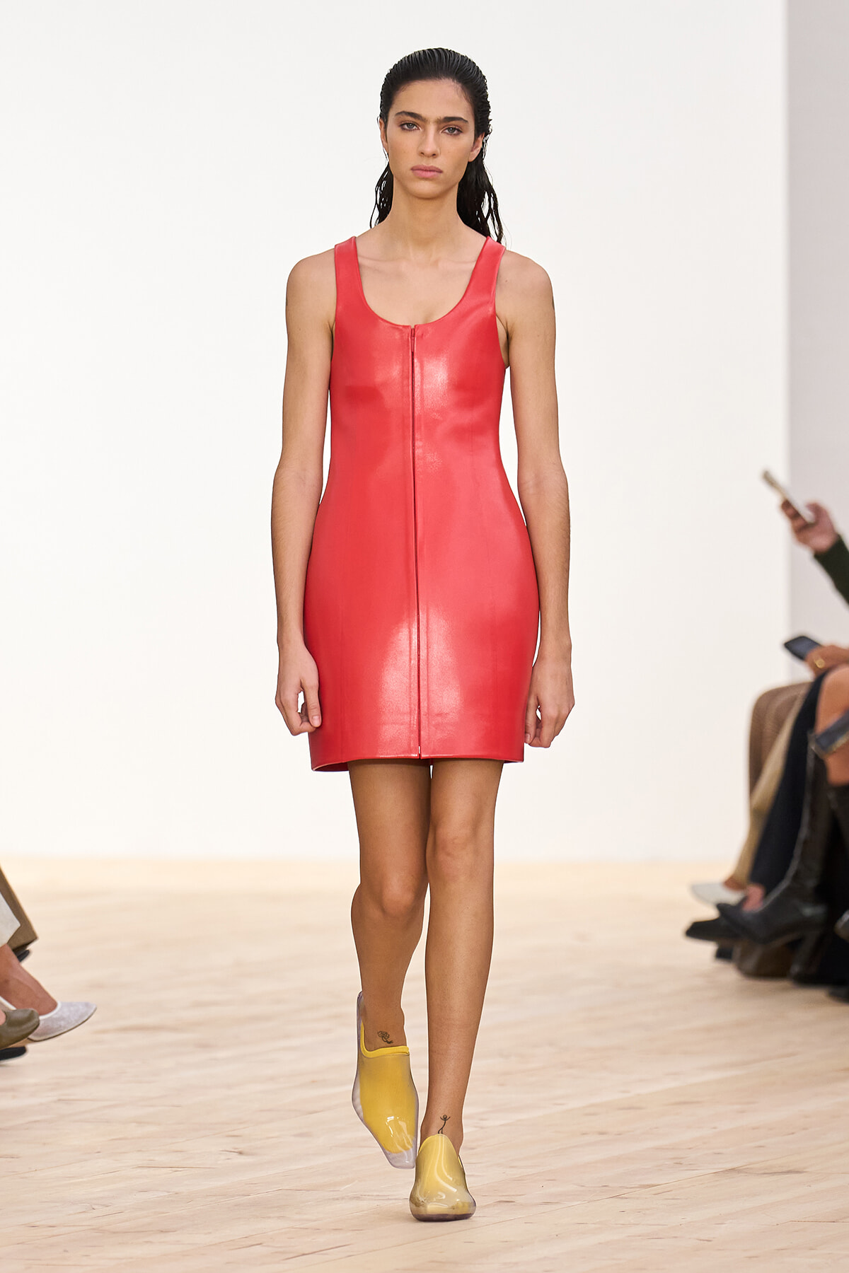 Model walking a runway in a sleeveless red leather mini dress with a centered vertical seam, paired with yellow slip-on shoes.