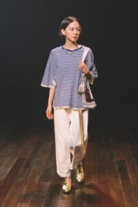 Young woman walks on a dark stage wearing a blue-and-white striped oversized top, white wide-leg pants, gold loafers, and a tote bag over her shoulder.