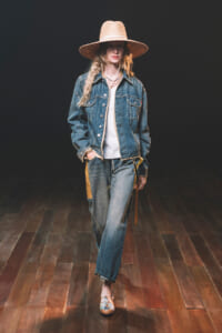 Model walking a runway in a denim jacket over a white top, wide-leg jeans with yellow side panels, and a large straw hat.