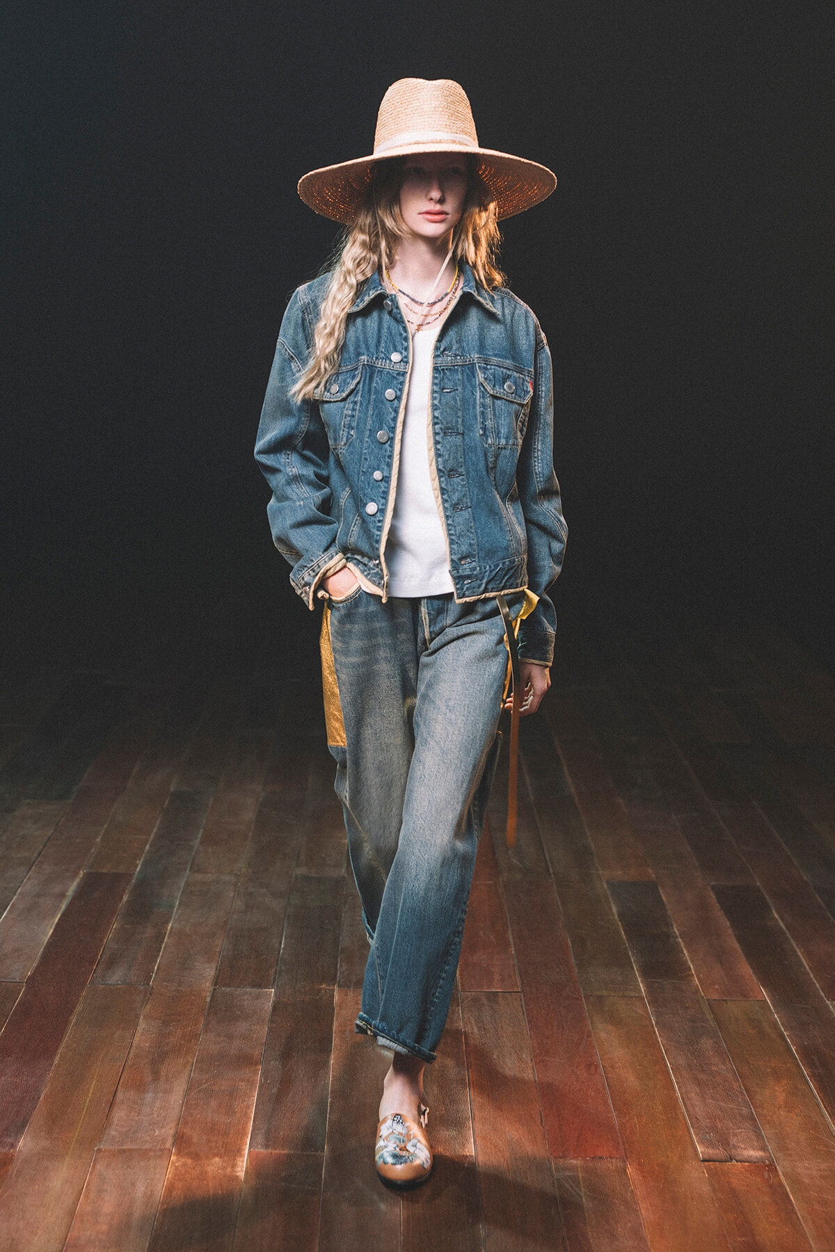 Model walking a runway in a denim jacket over a white top, wide-leg jeans with yellow side panels, and a large straw hat.