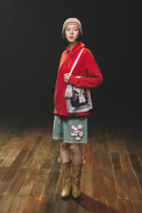 Young woman in a red oversized pullover, beige knit hat, and a green patchwork skirt with boots, carrying a white patterned tote bag, standing on a wooden floor against a dark background.