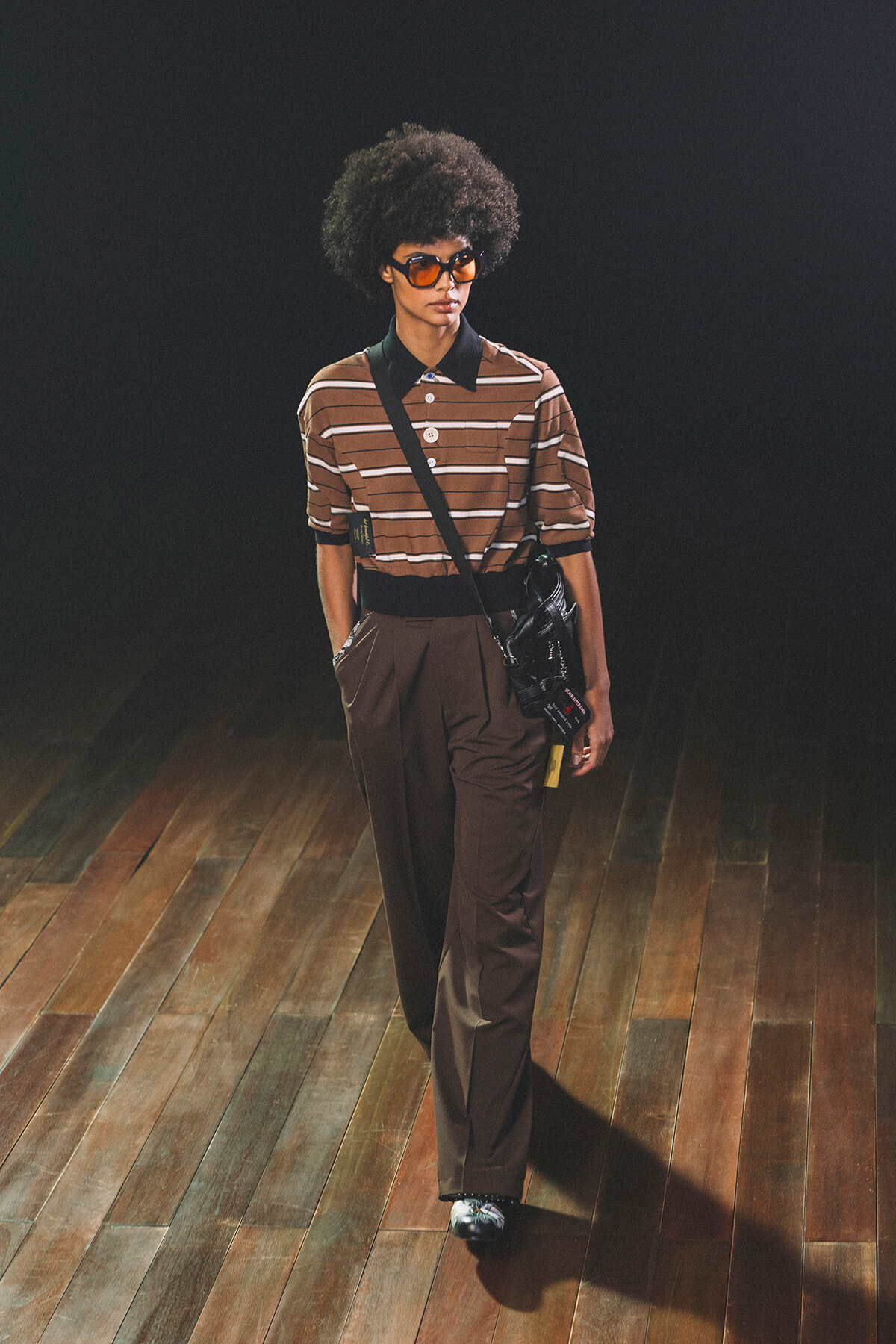 Model on a wooden runway wearing a brown striped polo, wide black belt, brown wide-leg pants, orange sunglasses, and a crossbody bag.