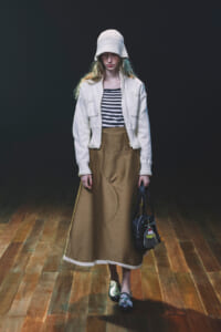 Model wearing a white knit hat, striped top, cream cardigan, and tan ankle-length skirt, holding a black handbag on a wooden floor with a dark backdrop.