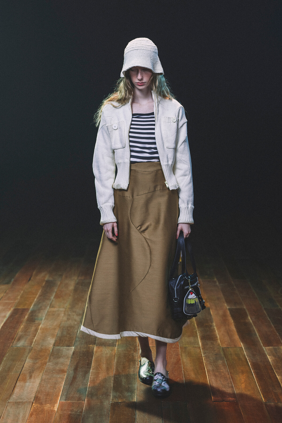 Model wearing a white knit hat, striped top, cream cardigan, and tan ankle-length skirt, holding a black handbag on a wooden floor with a dark backdrop.