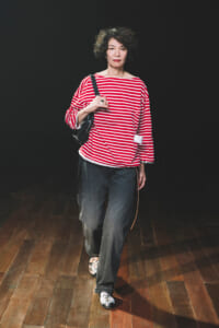 Older woman walking on a dark stage, wearing a red-and-white striped top, loose dark jeans, floral shoes, and a black shoulder bag.
