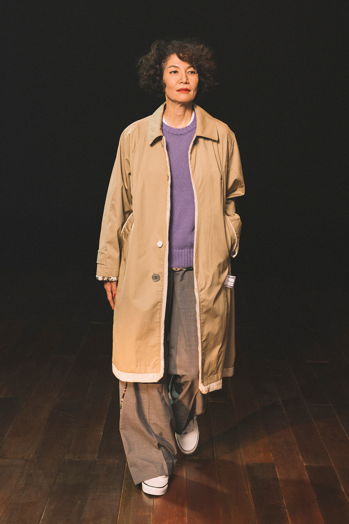 Model walking on a runway in a beige trench coat over a lavender sweater and gray wide-leg pants on a dark backdrop.
