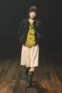 Young woman in a dark beanie and jacket over a mustard cardigan, a beige polka-dot midi skirt, and black embroidered cowboy boots, standing on a wooden floor against a dark backdrop.