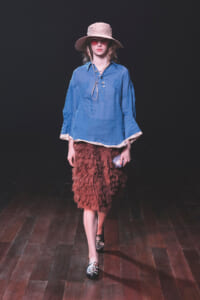 Model walking a runway in a blue long-sleeve blouse, brown textured knee-length skirt, pink sunhat, and pointed floral shoes; she wears red sunglasses and carries a small clutch.