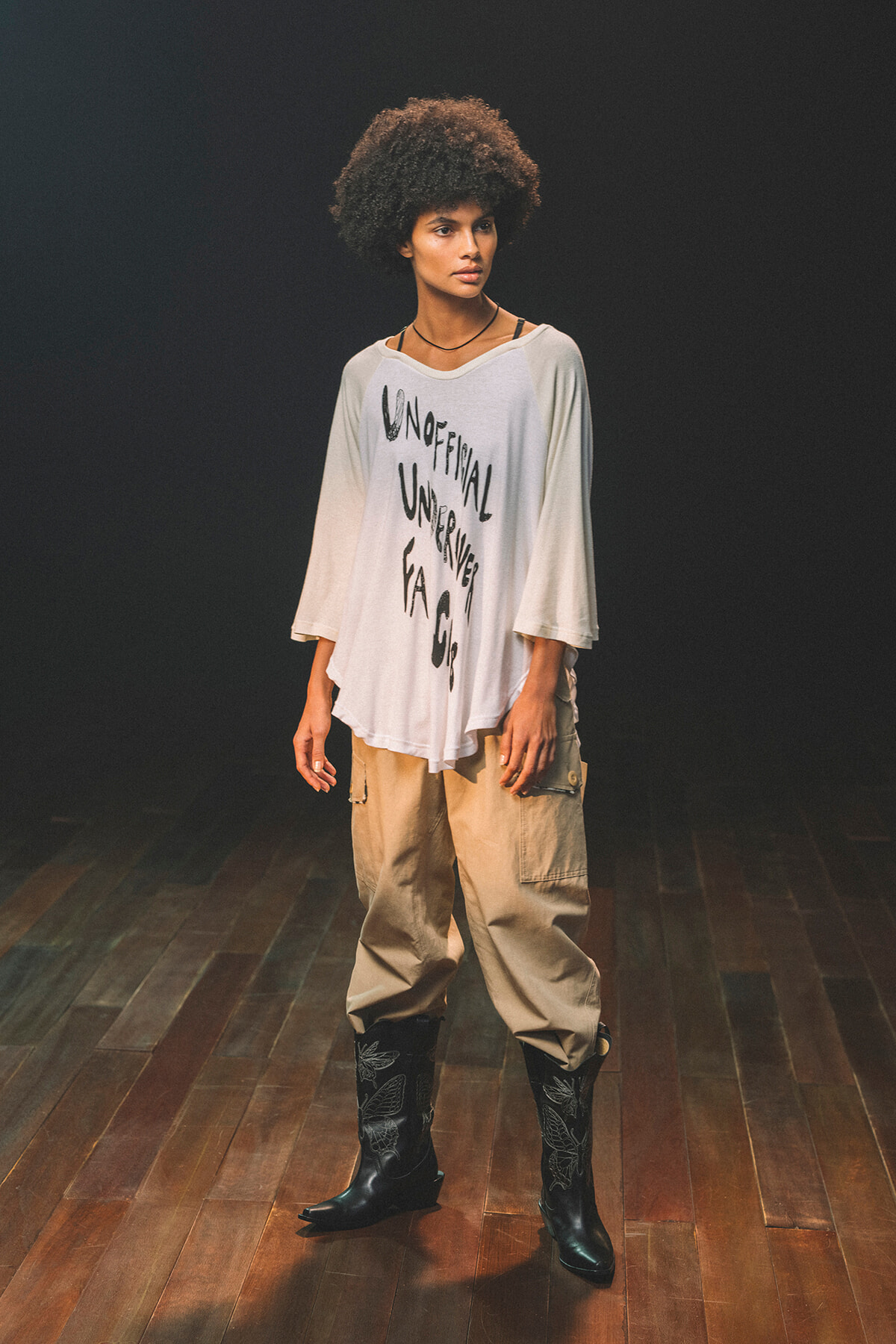 Model in a studio wearing an oversized white graphic tee, tan cargo pants, and black embroidered boots.
