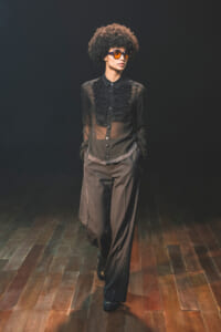 Model walks a dark runway in a sheer black blouse with ruffles, wide-leg pants, studded belt, and orange sunglasses.