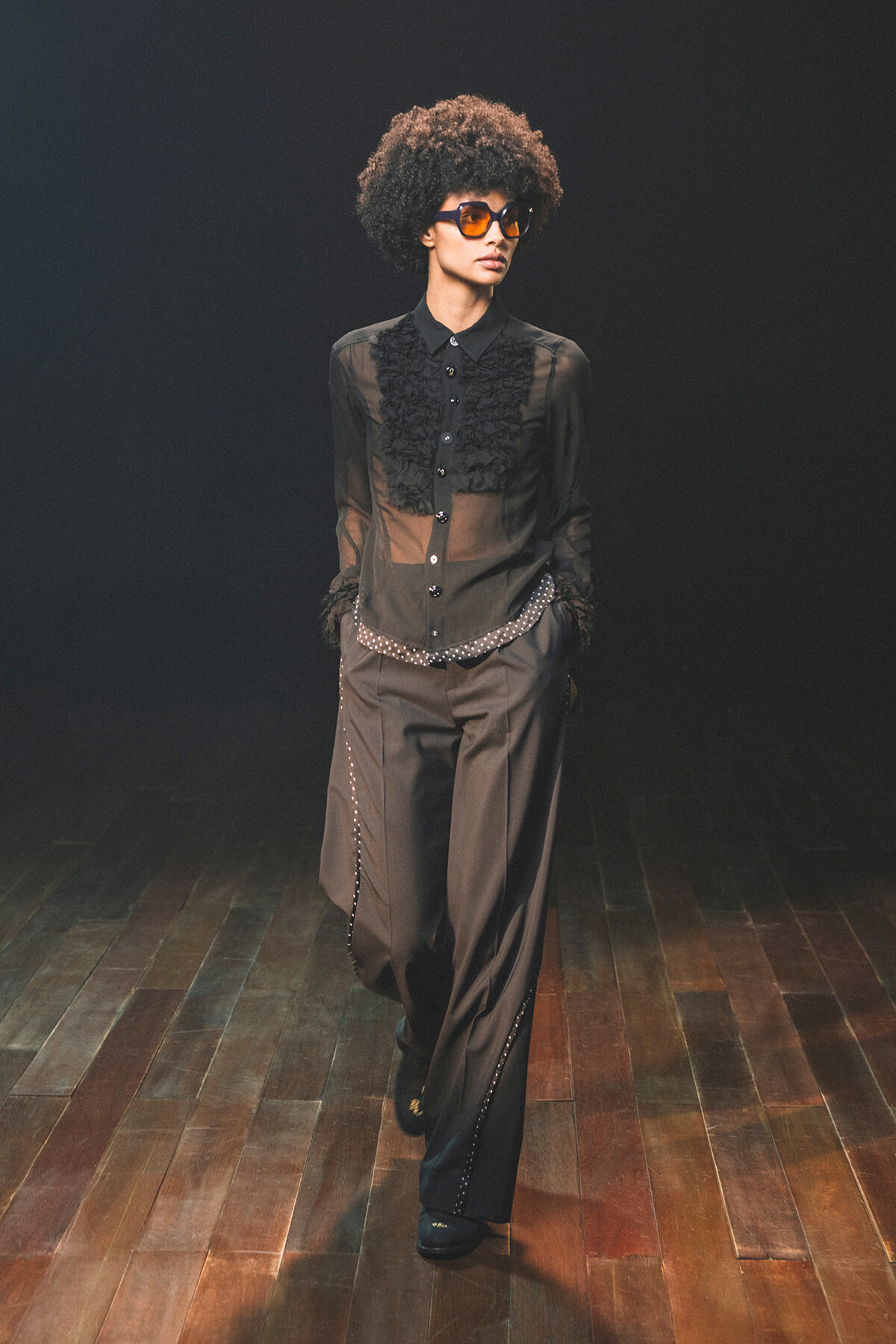 Model walks a dark runway in a sheer black blouse with ruffles, wide-leg pants, studded belt, and orange sunglasses.