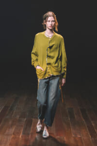 Model on a dark runway wearing an olive cardigan with front buttons and large pockets, paired with loose blue jeans and floral slippers on a wooden floor.