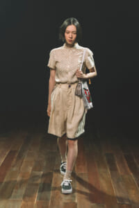 Woman in a beige polka-dot blouse and tan shorts with a crossbody bag, walking on a wooden stage against a dark background.