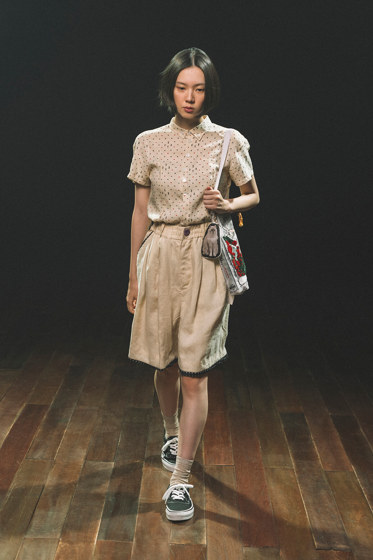 Woman in a beige polka-dot blouse and tan shorts with a crossbody bag, walking on a wooden stage against a dark background.