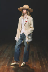 Model wearing a wide-brim straw hat, cream fringed blazer with pins, white top, jeans, and gold flats in a dark studio backdrop.