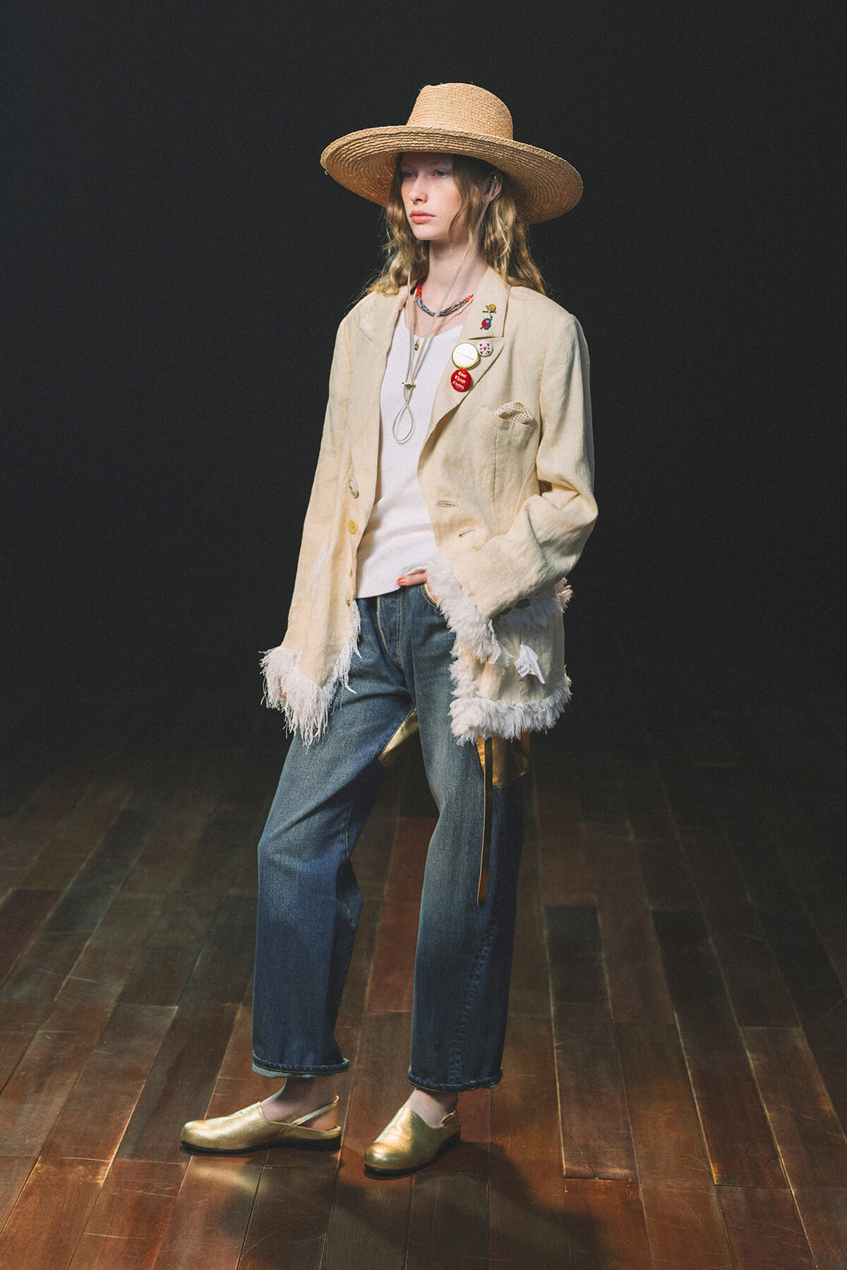 Model wearing a wide-brim straw hat, cream fringed blazer with pins, white top, jeans, and gold flats in a dark studio backdrop.