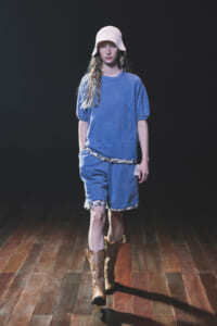 Model walks a runway in a blue loungewear set: oversized blue T-shirt, matching shorts with floral trim, white knit bucket hat, and brown cowboy boots, against a dark backdrop.