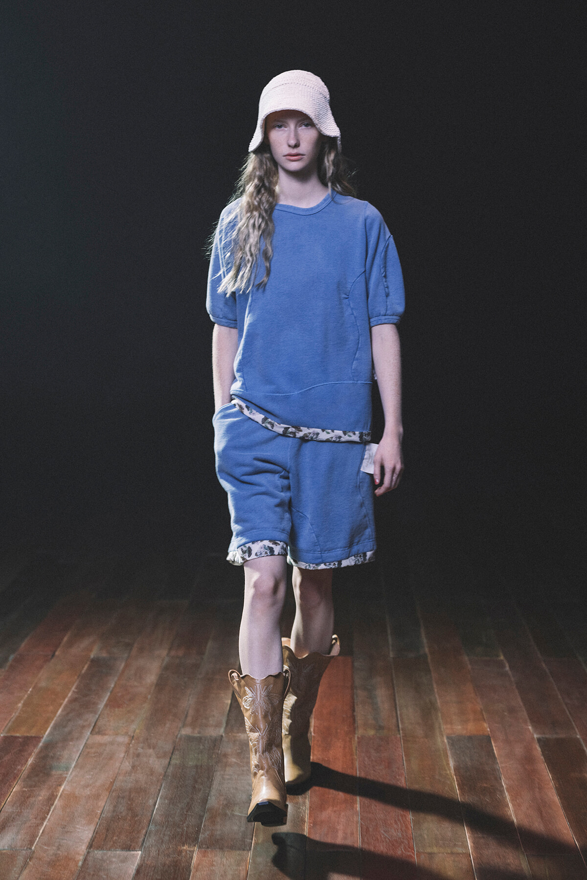 Model walks a runway in a blue loungewear set: oversized blue T-shirt, matching shorts with floral trim, white knit bucket hat, and brown cowboy boots, against a dark backdrop.