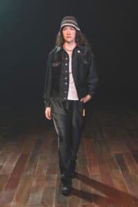Model walking on a dimly lit stage in a black denim jacket, white shirt, dark pants, and a striped bucket hat.