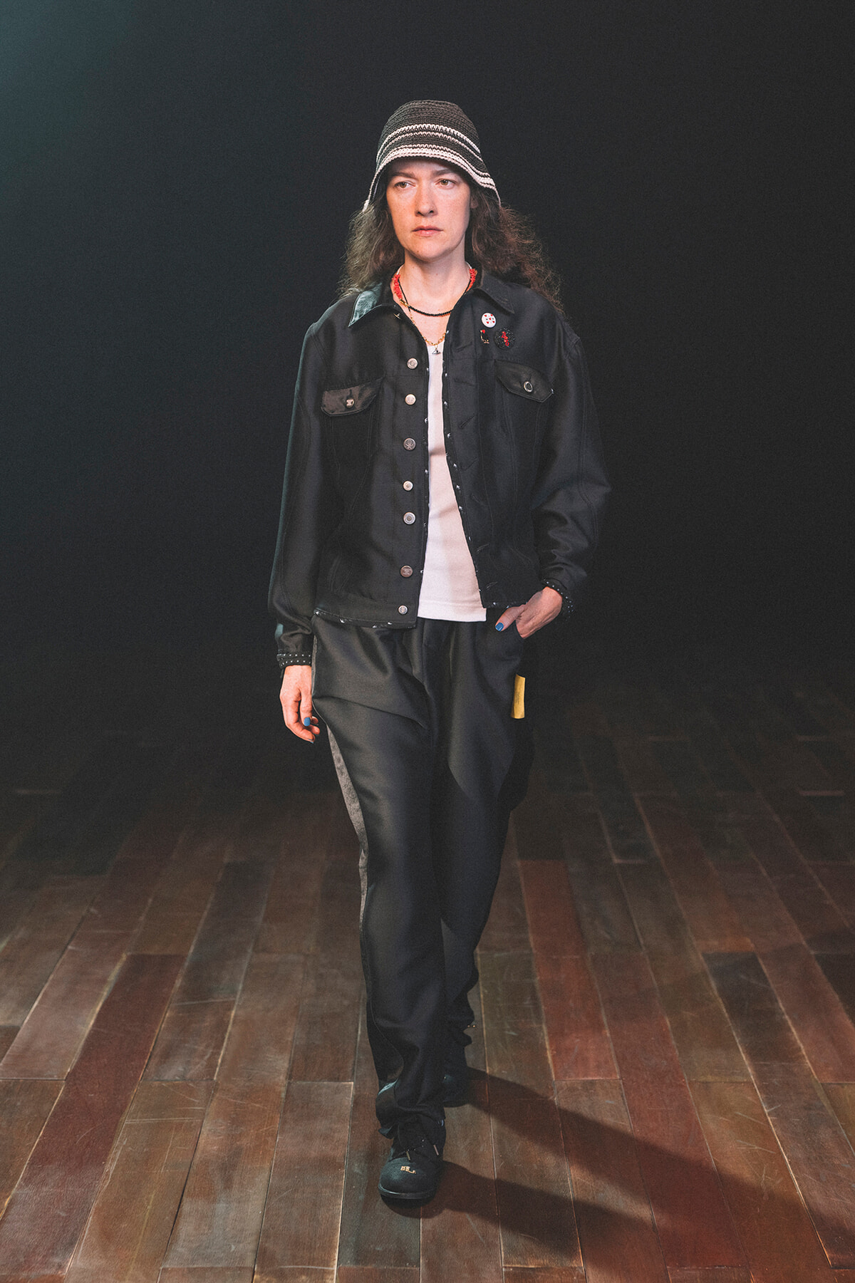 Model walking on a dimly lit stage in a black denim jacket, white shirt, dark pants, and a striped bucket hat.