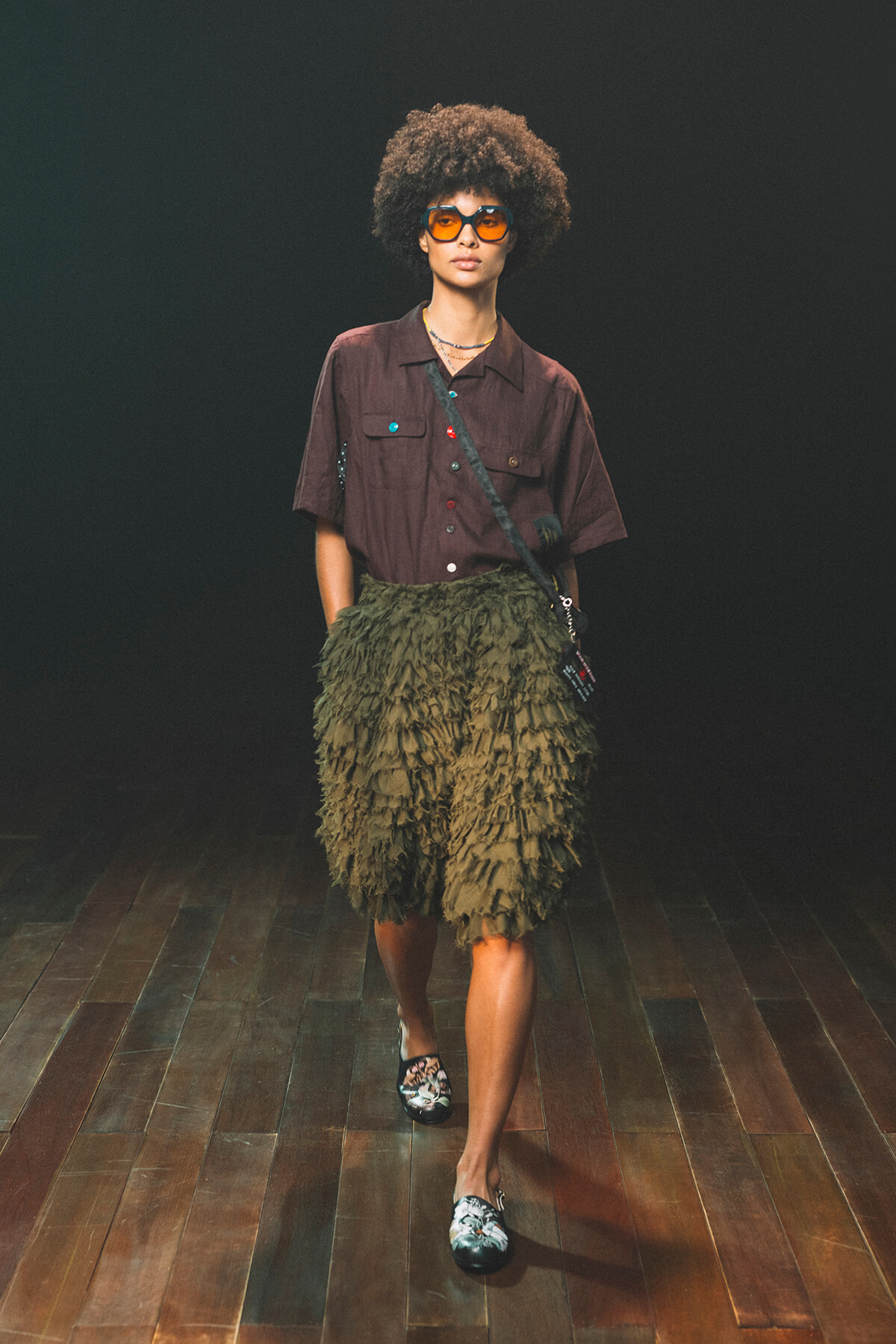 Model on a runway wearing a brown short-sleeve shirt and olive-green textured ruffle skirt, with orange sunglasses and a crossbody bag.