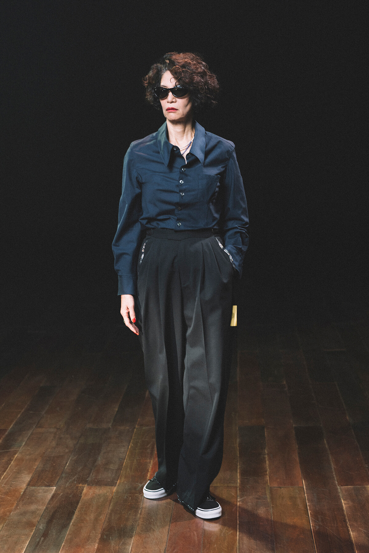 Person wearing a navy button-down shirt and wide black trousers, with sunglasses, standing on a wooden stage against a dark background.