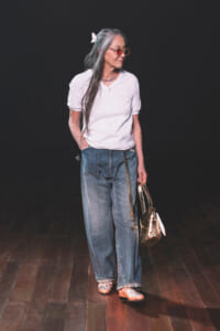 Older woman with gray hair, white top, loose jeans, butterfly hair clip, pink sunglasses, carrying a metallic handbag on a dark stage.